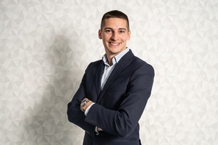 A confident young man in a navy blue suit and light blue dress shirt stands with his arms crossed, smiling at the camera. He is positioned against a geometric-patterned white background, conveying professionalism and approachability. This image represents career potential with a Master's in Management.