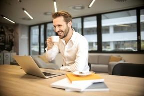 Smiling professional man in business attire drinking coffee while working on a laptop in a modern office, symbolizing career opportunities with a business management degree.