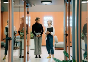 Two professionals conversing in a modern office corridor with glass walls, reflecting a collaborative and contemporary workplace environment.