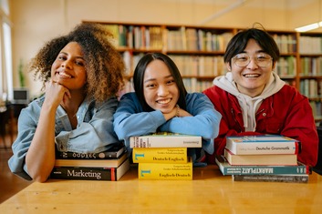 Three students sit at a wooden library table, smiling and leaning on stacks of books including “Marketing,” with bookshelves in the background.