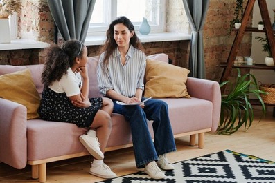 A school counselor sits on a pink sofa holding a clipboard, listening as a young girl beside her talks, in a bright room with a brick wall, large windows and houseplants.